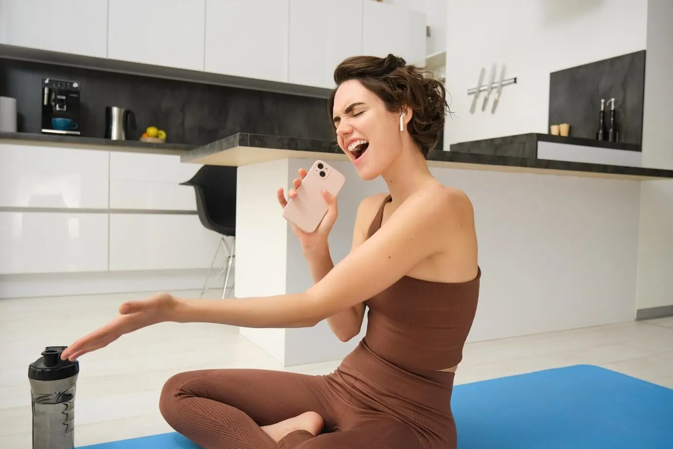 A happy young brunette fitness girl sits on a yoga mat on the kitchen floor with a water bottle, working out from home.